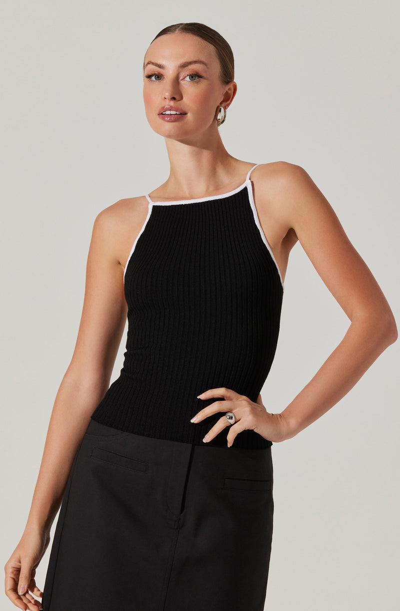 A woman is wearing a sleeveless black ribbed top with white trim along the neckline and straps. She pairs it with a high-waisted black skirt that has a clean, straight silhouette and a front slit. The outfit has a minimalist, chic style.