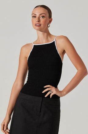 A woman is wearing a sleeveless black ribbed top with white trim along the neckline and straps. She pairs it with a high-waisted black skirt that has a clean, straight silhouette and a front slit. The outfit has a minimalist, chic style.