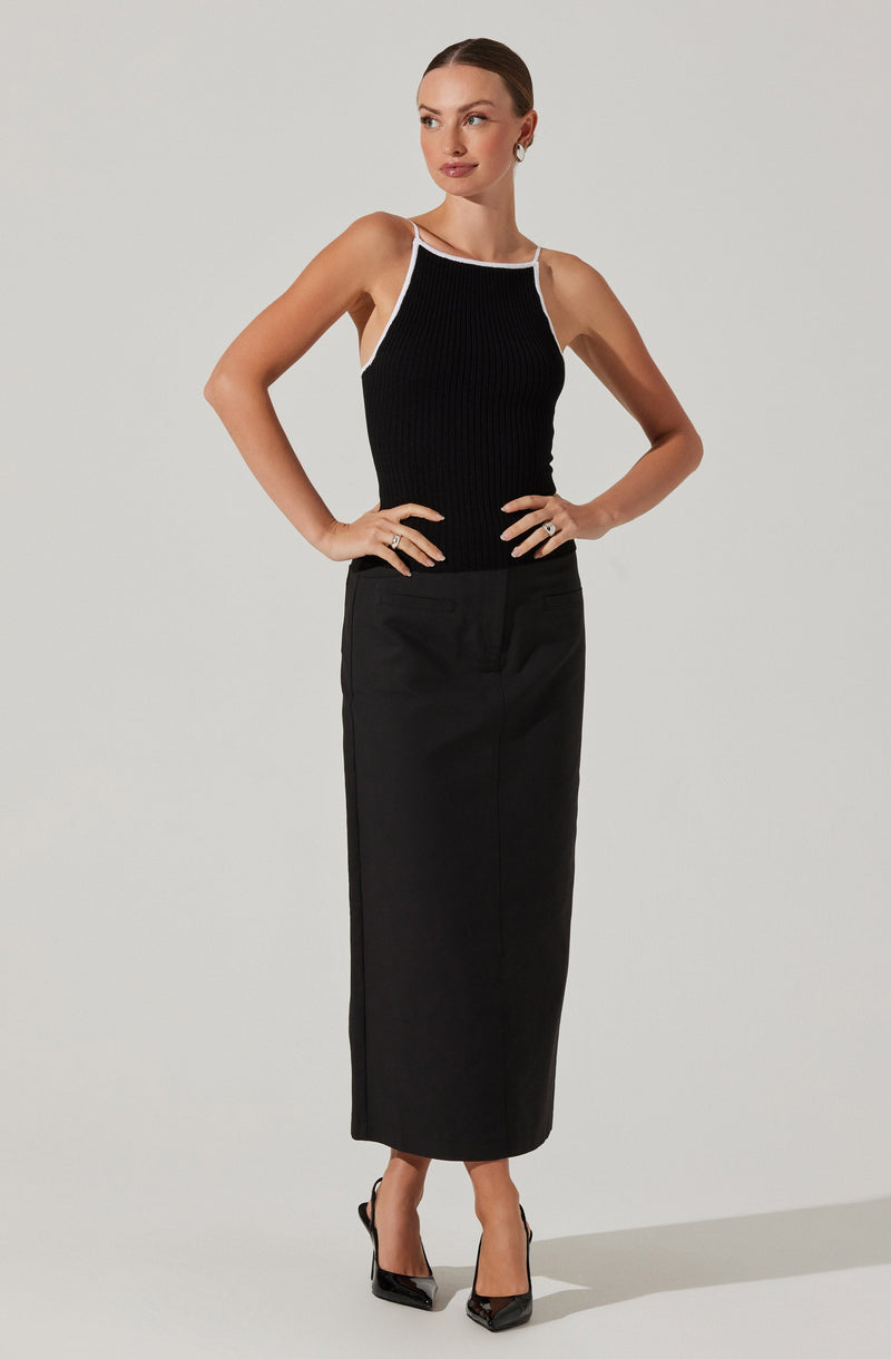 A woman is wearing a sleeveless, black ribbed top with white trim around the neckline and straps. She pairs it with a high-waisted, black, ankle-length skirt. She is also wearing black high-heeled shoes.