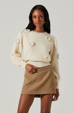 A woman is wearing a cream-colored, fuzzy sweater with textured, fluffy circular embellishments scattered across the front and sleeves. She is also wearing a high-waisted, camel-colored, A-line mini skirt made of a soft, wool-like material.