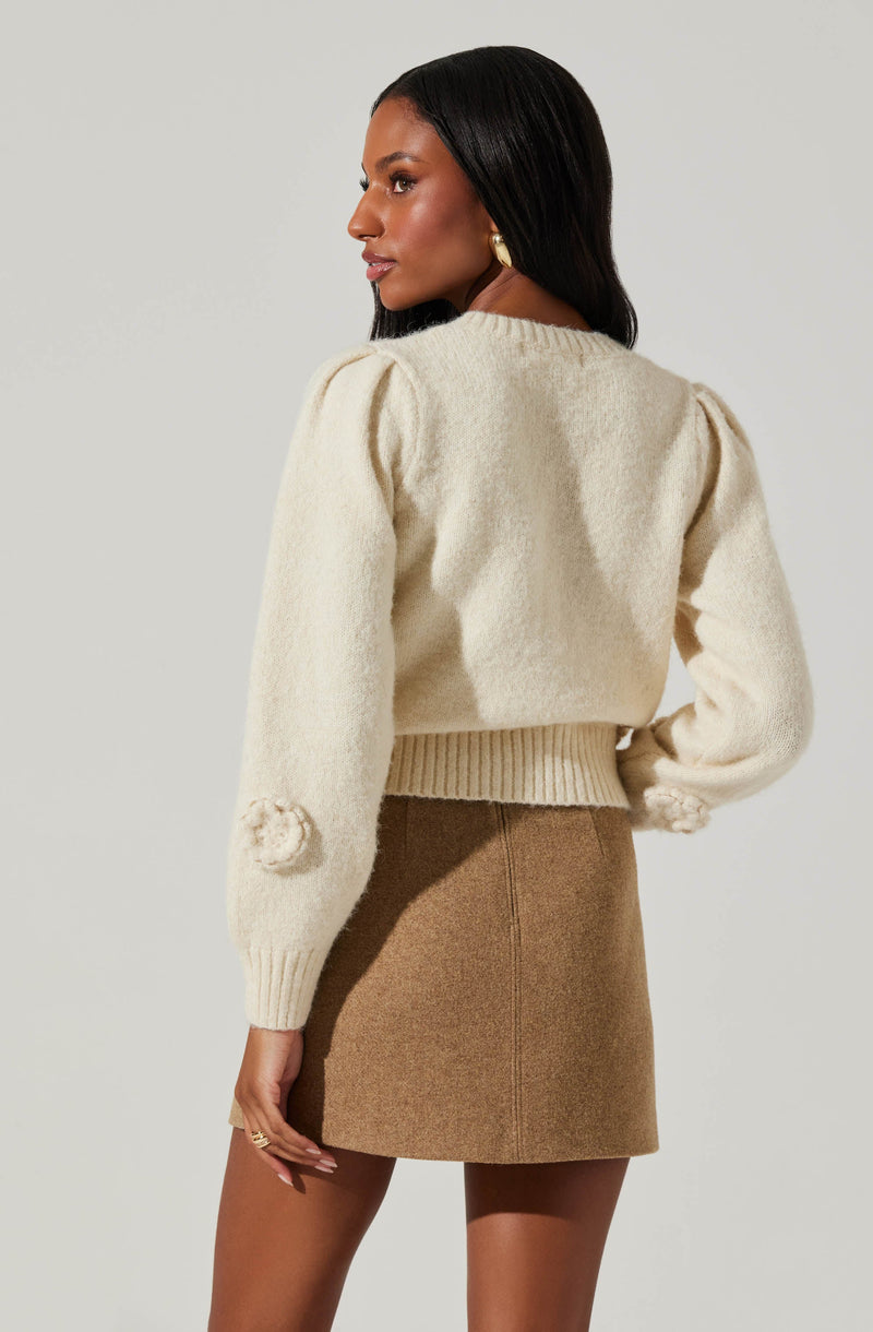 A woman is wearing a cream-colored, long-sleeved knit sweater with subtle puffed shoulders and decorative pom-poms on the sleeves. She pairs it with a high-waisted, camel-colored mini skirt made of a textured fabric.
