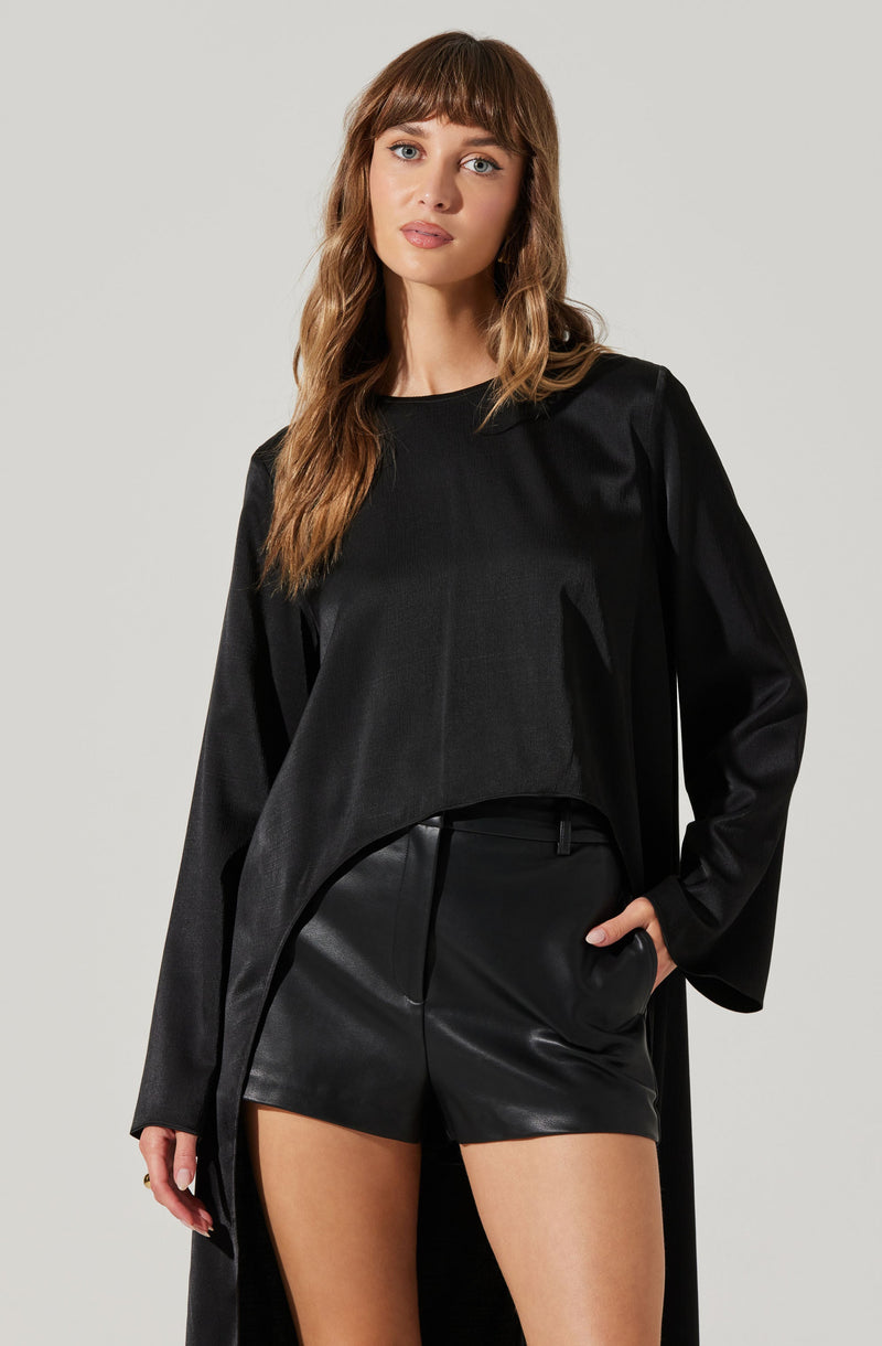 A woman is wearing a black long-sleeve top with a loose fit and subtle pleats on the front, paired with high-waisted black leather shorts.