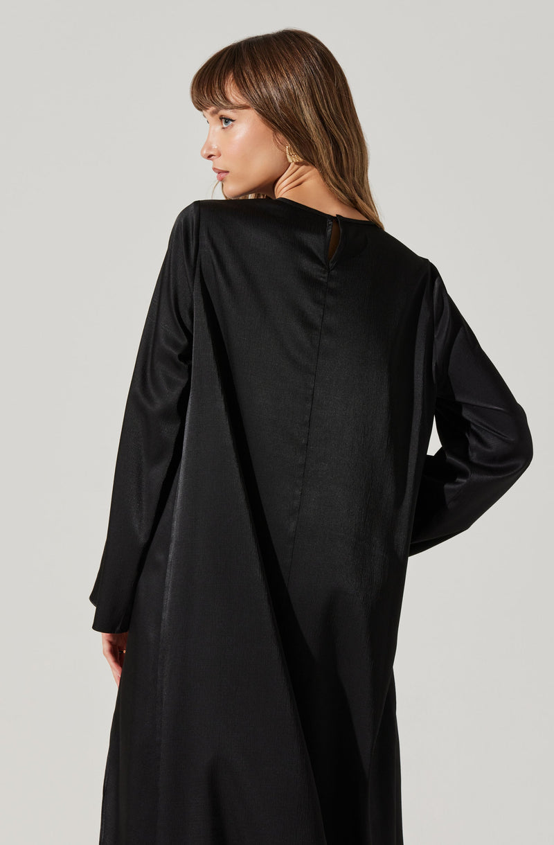 A woman is wearing a long, black, satin-like dress or coat with a high neckline and long sleeves. The back of the garment features a small keyhole opening at the neck, fastened with a button. The fabric has a smooth and slightly shiny texture.