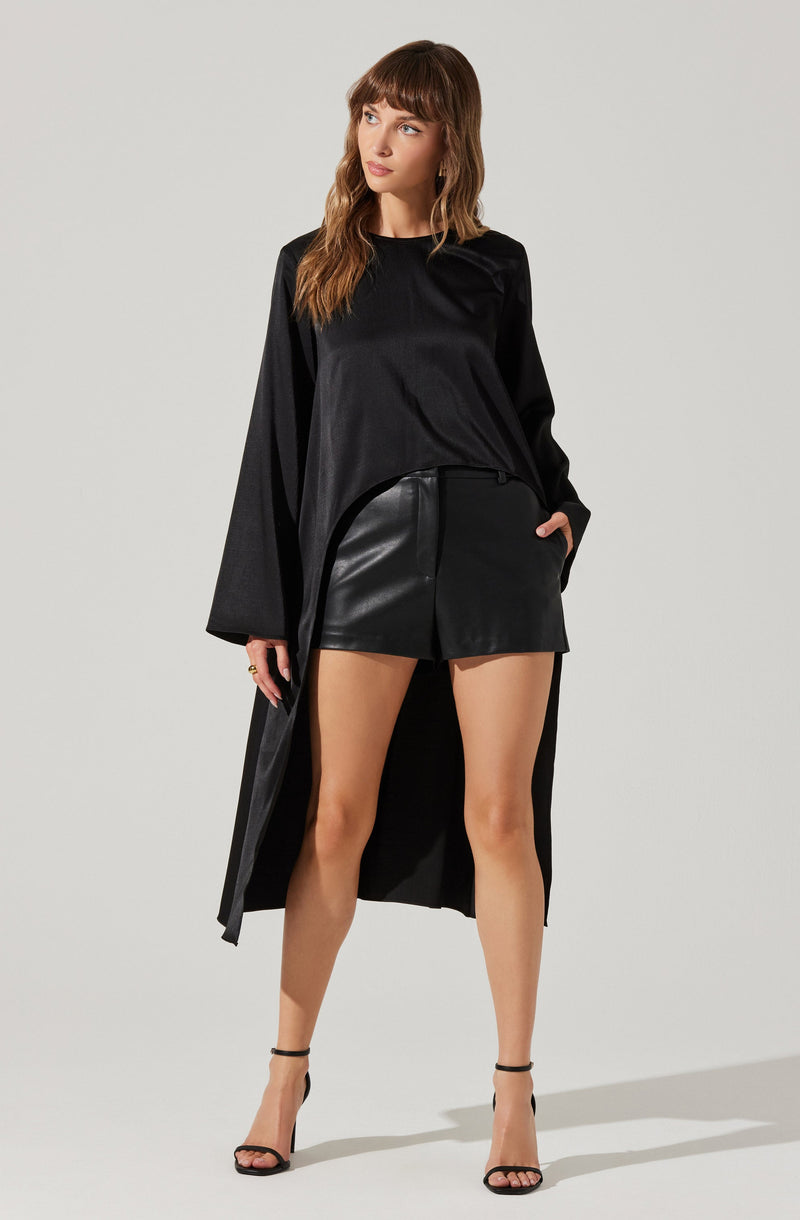 A woman is wearing a black long-sleeve top with a shiny, silky texture, paired with black faux leather shorts. She has on black strappy high-heeled sandals. She is also wearing a long black coat that drapes down to her calves. Her hair is brown, wavy, and shoulder-length.