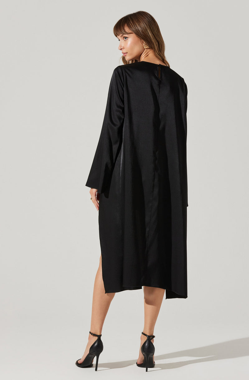 A woman is wearing a long, black satin dress with a shiny finish. The dress features long sleeves and a slit on the side, revealing her legs. The back of the dress has a vertical seam running down the center. She pairs the dress with black high-heeled sandals that have ankle straps. Her hair is shoulder-length and brown, styled with soft waves.