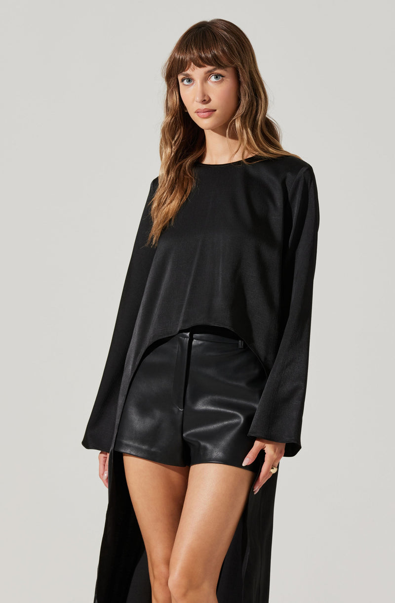 A woman is wearing a black long-sleeve top with a high neckline and slightly flared sleeves. She pairs it with high-waisted black leather shorts. The outfit creates a sleek, monochromatic look.
