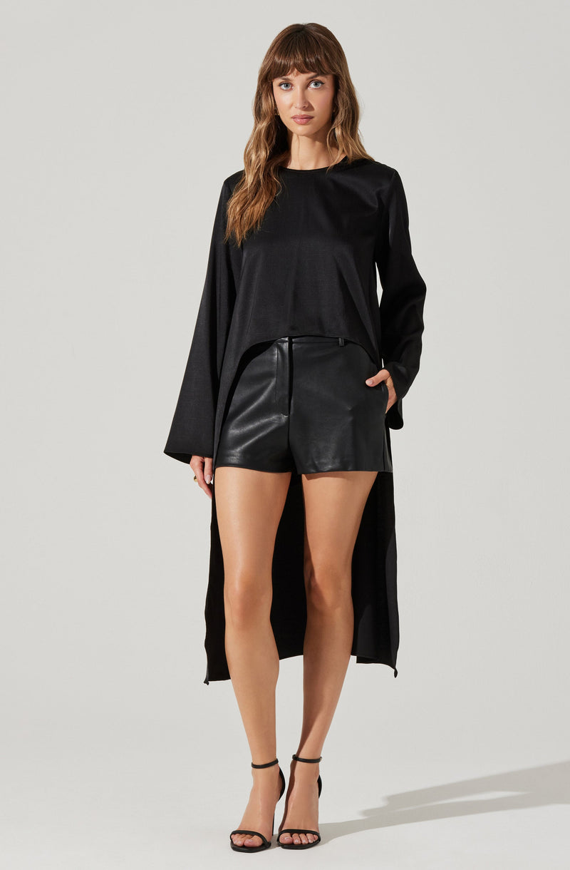 A woman is wearing a long black coat with wide sleeves, a black long-sleeve top, and high-waisted black leather shorts. She has on black strappy high-heeled sandals and has her hand in one of the shorts' pockets.