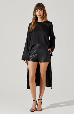 A woman is wearing a long black coat with wide sleeves, a black long-sleeve top, and high-waisted black leather shorts. She has on black strappy high-heeled sandals and has her hand in one of the shorts' pockets.