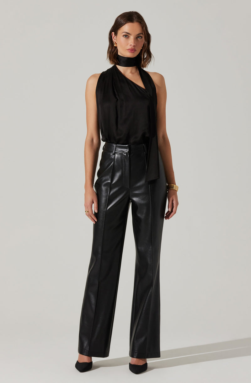 A woman is wearing a sleeveless, black satin top with a loose fit and a high neckline that drapes softly. She pairs it with high-waisted, black leather wide-leg pants. She accessorizes with a black choker around her neck, small hoop earrings, a gold watch on her left wrist, and black pointed-toe heels.