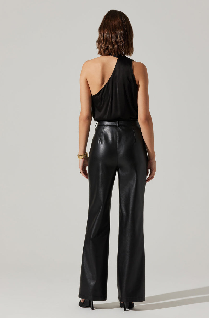 A woman is wearing a black sleeveless top with a one-shoulder design, featuring a draped, loose fit. She pairs it with high-waisted black leather wide-leg pants. She accessorizes with a gold bracelet on her left wrist and black high-heeled shoes.
