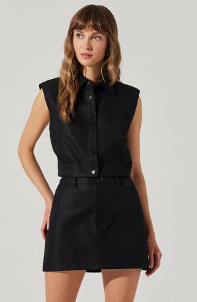 A woman is wearing a sleeveless black button-up shirt with a collar, paired with a black high-waisted skirt. The shirt has shoulder pads, giving it a structured appearance.