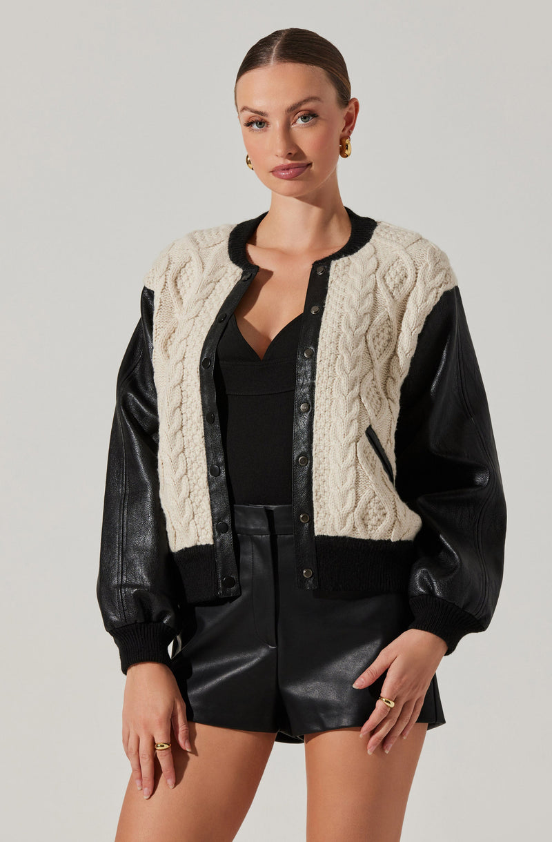 A young woman is wearing a varsity-style jacket with black leather sleeves, black collar, cuffs, and waistband, and a white cable-knit body. Underneath, she has on a black top, and she is also wearing black leather shorts. She accessorizes with gold earrings and rings.