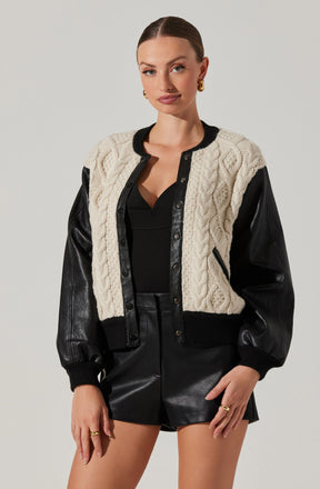 A young woman is wearing a varsity-style jacket with black leather sleeves, black collar, cuffs, and waistband, and a white cable-knit body. Underneath, she has on a black top, and she is also wearing black leather shorts. She accessorizes with gold earrings and rings.