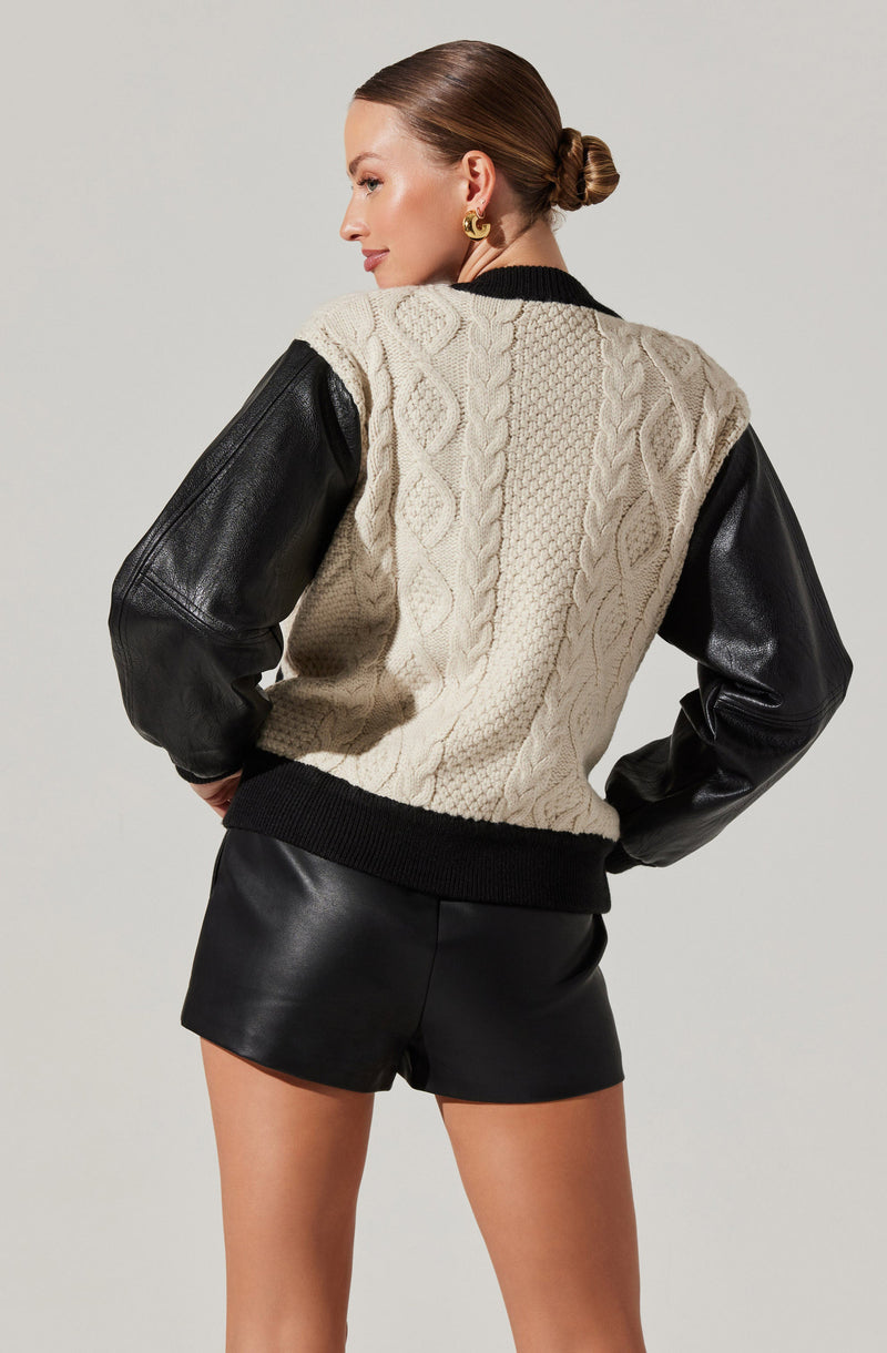 A woman is wearing a varsity-style sweater with a cream-colored knitted front featuring cable patterns and black ribbed cuffs, collar, and hem. The sweater has black faux leather sleeves. She is also wearing black leather shorts.