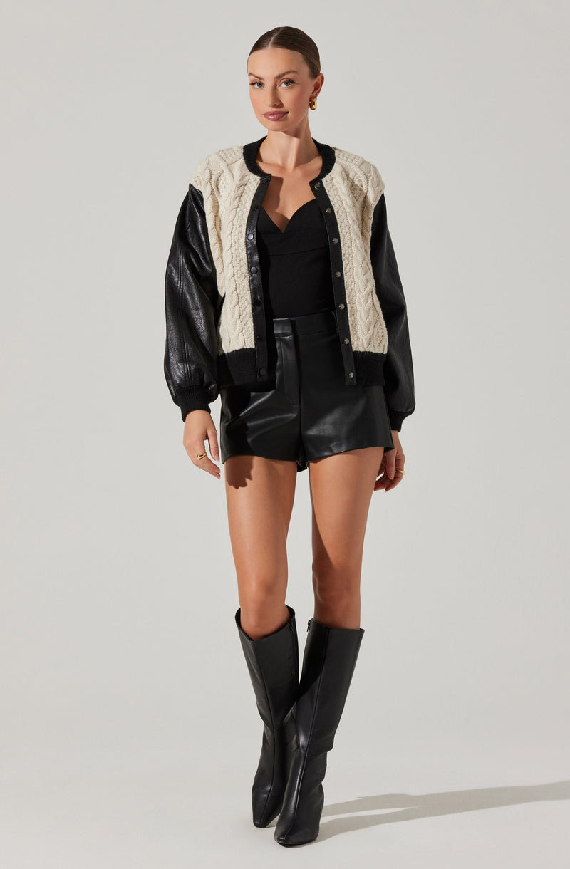 A woman is wearing a varsity-style jacket with a cream cable knit body and black leather sleeves, paired with a black top, black leather shorts, and tall black leather boots.