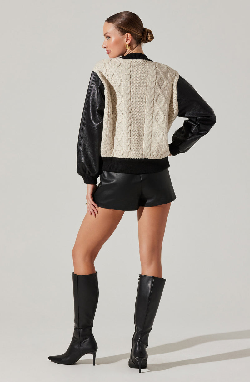A woman is wearing a cropped, sleeveless, cable-knit sweater vest in a cream color, layered over a black long-sleeve top with shiny leather sleeves. She pairs this with black leather shorts and black knee-high heeled boots.