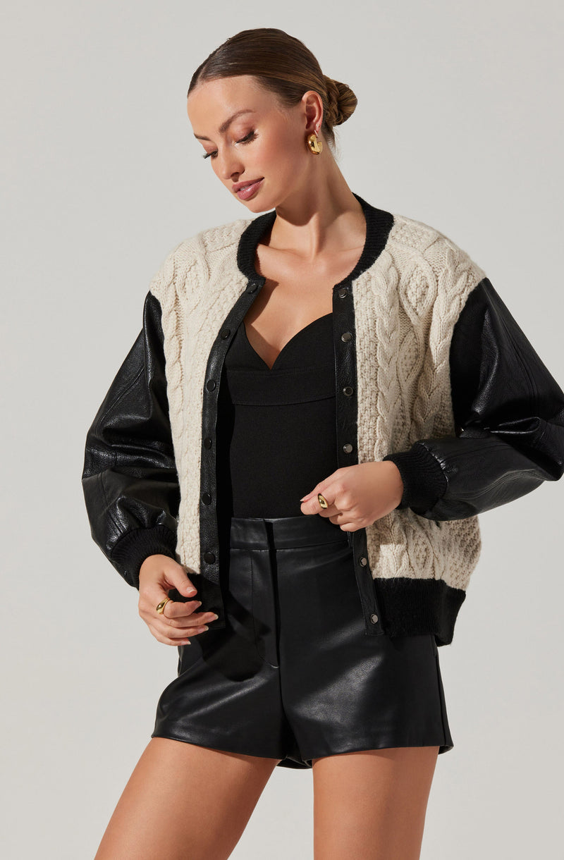 A woman is wearing a varsity-style jacket with a knitted cream-colored front featuring cable knit patterns, black ribbed collar and cuffs, and black faux leather sleeves. She is also dressed in a black V-neck top and black faux leather shorts.
