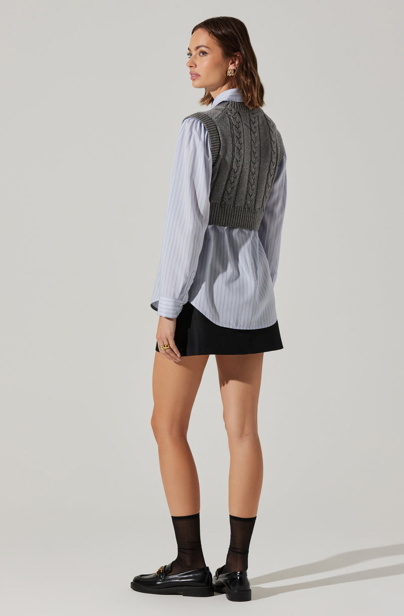 A woman is wearing a light blue, long-sleeved, button-down shirt with vertical white stripes. Over the shirt, she has on a dark gray, ribbed, sleeveless knitted vest with cable knit pattern. She is also wearing a short black skirt, black ankle socks, and black loafers with gold buckle accents.