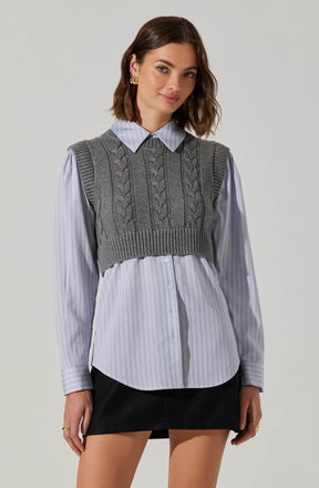 A young woman is wearing a light blue collared shirt with thin vertical white stripes, buttoned down the front. Over the shirt, she has on a gray knit sleeveless sweater vest featuring braided cable patterns on the front and ribbed details at the hem and armholes. She pairs the top with a black skirt.