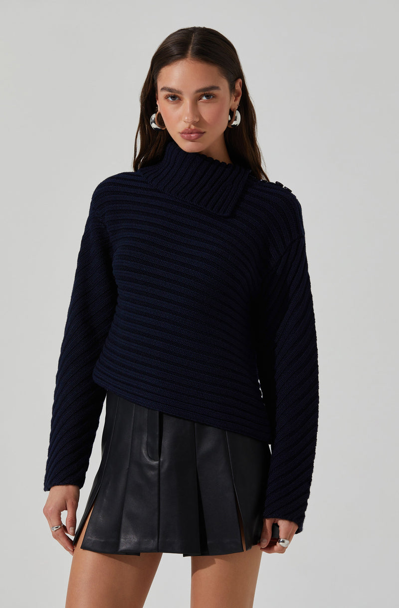 A young woman is wearing a dark navy blue, chunky knit sweater with a wide, ribbed turtleneck that extends across one shoulder. She pairs it with a black, high-waisted leather mini skirt featuring front pleats and side zipper details. She accessorizes with silver hoop earrings and rings.