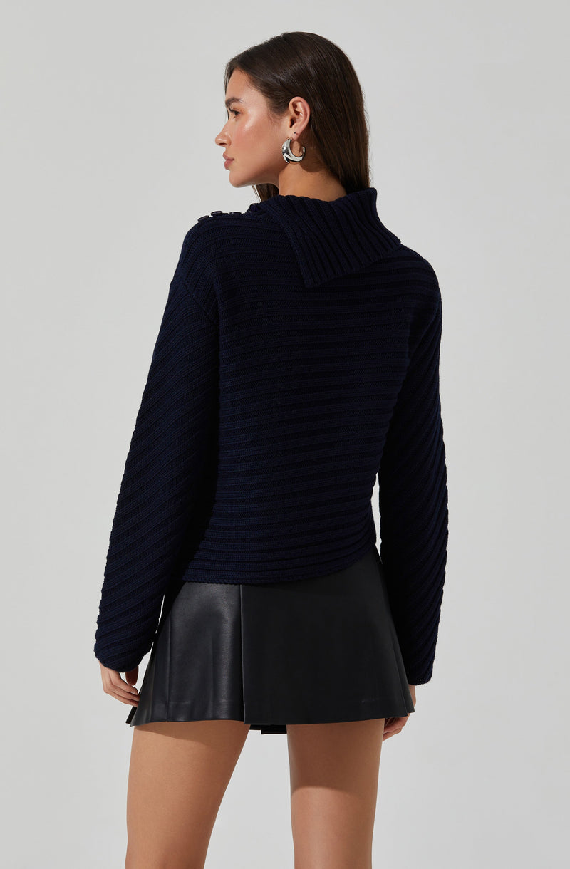 A woman is wearing a dark navy blue, chunky, ribbed knit sweater with a large, folded collar, paired with a black leather mini skirt. She has her dark brown hair parted to the side and is accessorized with hoop earrings.