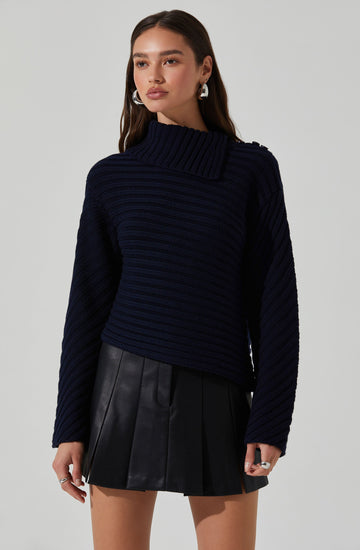 A woman is wearing a navy blue, chunky knit sweater with a high, wide collar that covers part of her neck. The sweater has a textured, ribbed pattern with horizontal stripes. She pairs it with a black leather mini skirt that features pleats. She is accessorized with large silver hoop earrings and rings on her fingers.