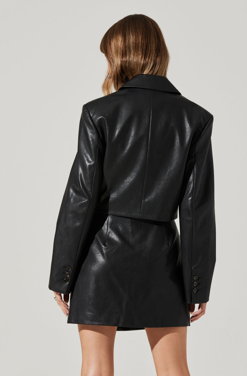 A woman is wearing a black leather blazer with long sleeves and a notch lapel collar. The blazer has four buttons on each sleeve cuff. She is also wearing a matching black leather skirt that is above knee length.
