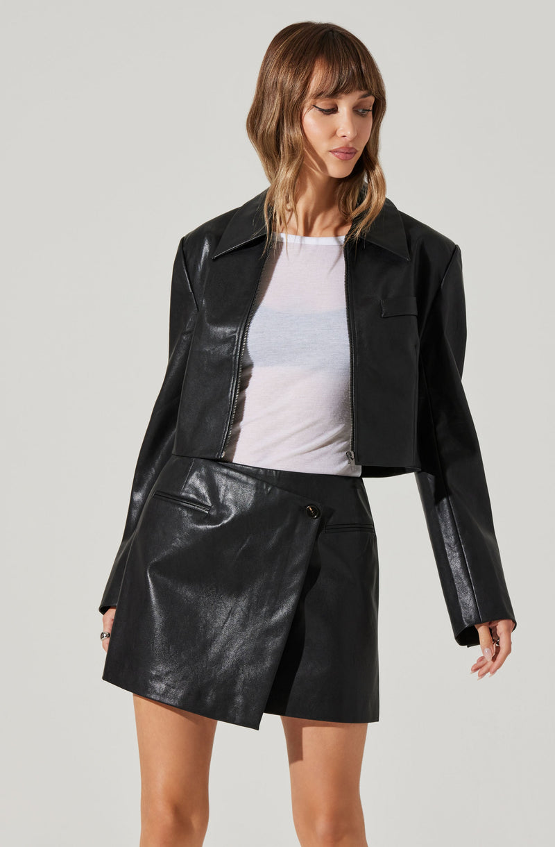 A woman is wearing a black cropped leather jacket with a collar and long sleeves. She has on a white T-shirt underneath and a high-waisted black leather mini skirt with a side snap closure.