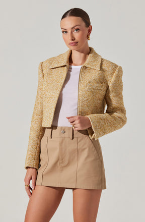 A woman is wearing a light yellow and beige textured cropped jacket with a collar and front zipper, paired with a beige high-waisted mini skirt that has front pleats and side seam pockets. She also has on a white top underneath the jacket.