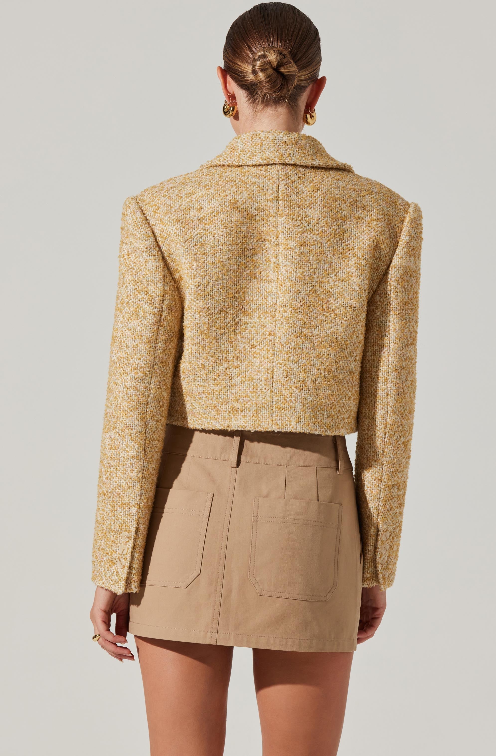 A woman is wearing a cropped, textured, yellowish-tan blazer with long sleeves and a collar. She pairs it with a high-waisted beige mini skirt that has two back pockets. The overall look is stylish and polished.