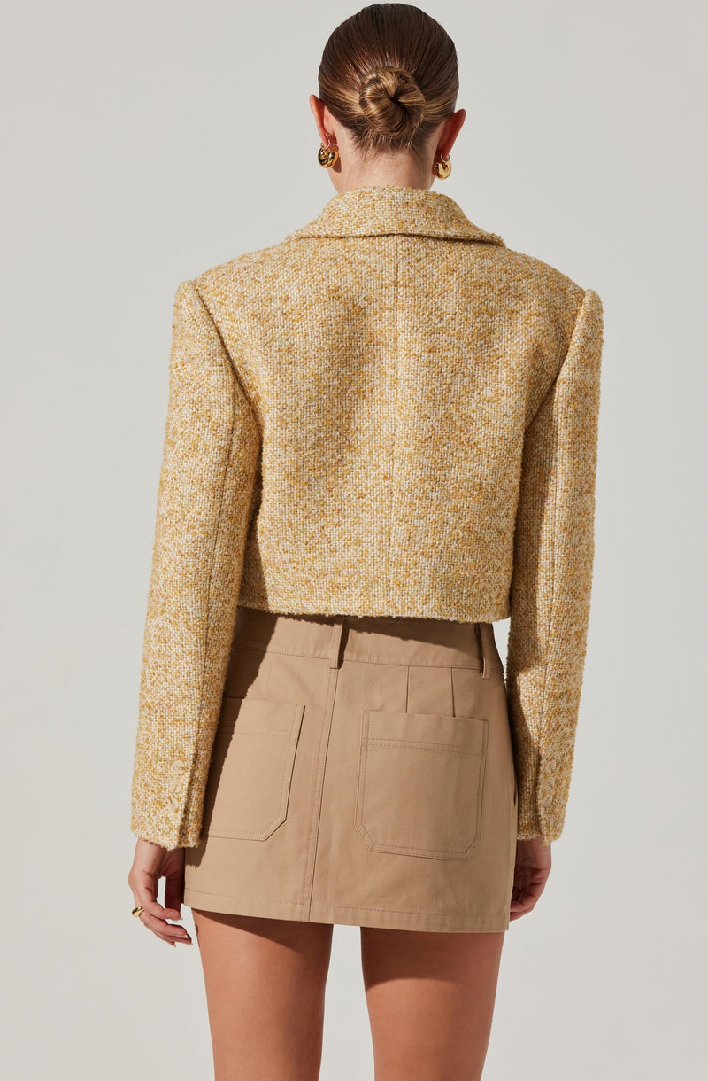 A woman is wearing a cropped, textured, yellowish-tan blazer with long sleeves and a collar. She pairs it with a high-waisted beige mini skirt that has two back pockets. The overall look is stylish and polished.