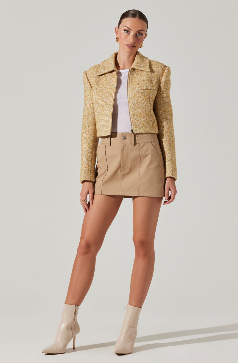 The woman is wearing a light beige, textured cropped jacket with a collar and long sleeves. Underneath, she has a white top. She is also wearing a high-waisted beige mini skirt with vertical seam details and belt loops. Her footwear consists of sleek, pointed-toe ankle boots in a shiny, light beige or cream color with stiletto heels.