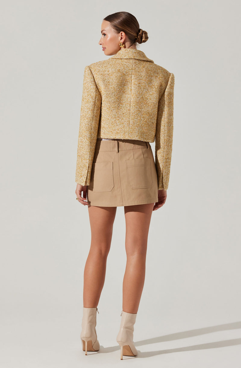 A woman is wearing a cropped, textured, multicolored blazer in shades of yellow and beige, with broad shoulders. She pairs it with a high-waisted beige mini skirt that features two back pockets. She completes the outfit with nude-colored ankle boots with high heels.