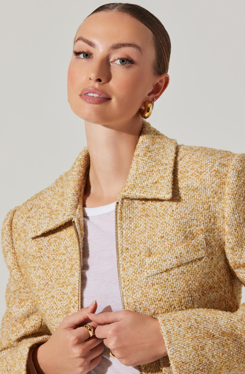 A woman is wearing a textured, multicolored tweed jacket in shades of yellow, orange, and cream. The jacket has a collar and a front zipper. Underneath, she has on a white T-shirt. She is accessorized with large, gold hoop earrings and gold rings on her fingers.