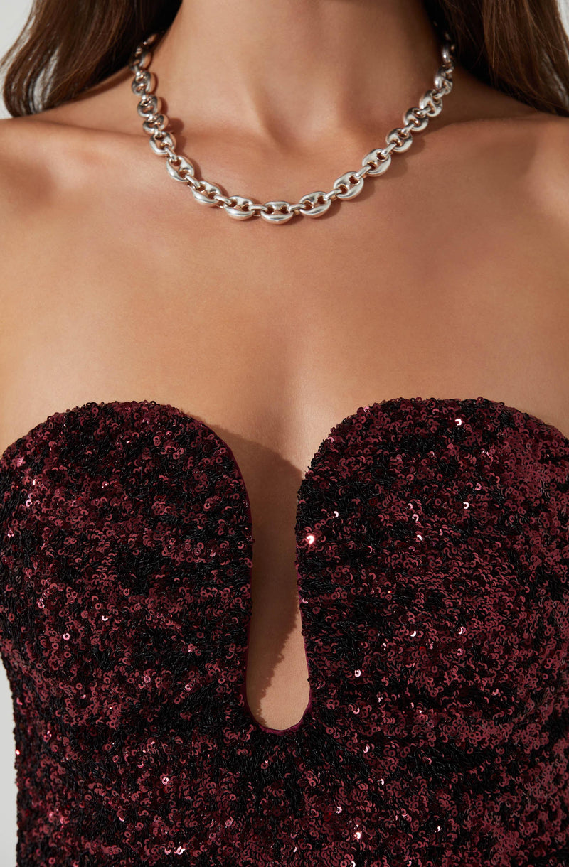 A woman is wearing a strapless dress covered in dark red sequins, with a deep sweetheart neckline that features a small V-shaped cut in the center. She has a chunky, silver chain necklace with large, oval links resting around her neck.