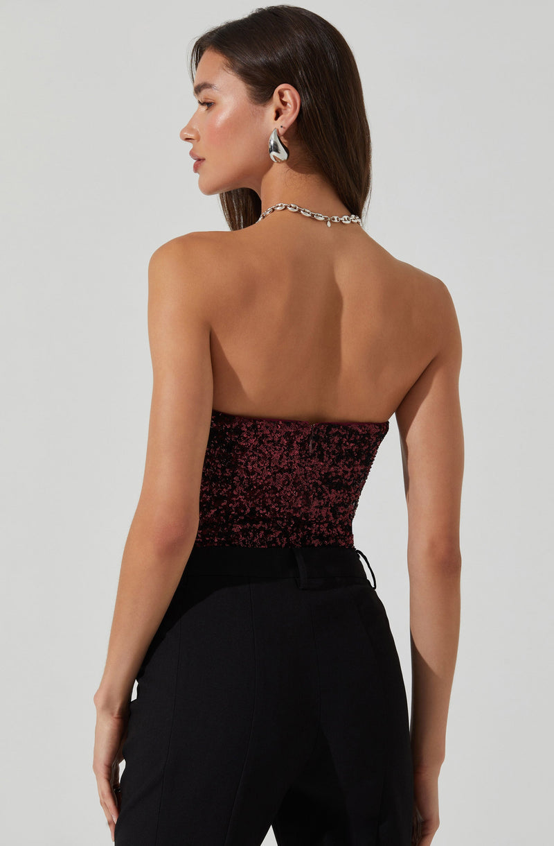 A woman is wearing a strapless top that features a fitted silhouette and is covered in black and red glitter. She pairs it with high-waisted black trousers. She accessorizes with large, asymmetric silver earrings and a chunky silver chain necklace.