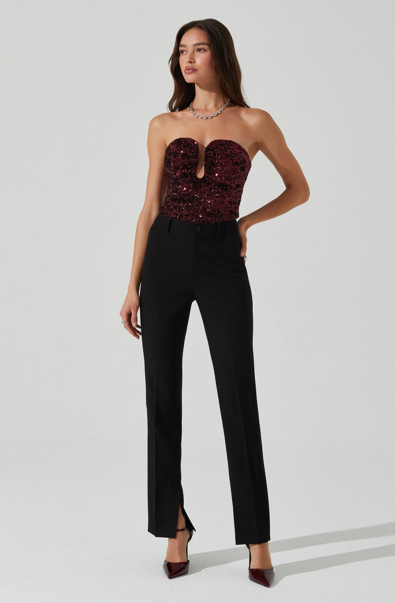 A woman is wearing a strapless, fitted top covered in dark red sequins with a central keyhole cutout. She pairs it with high-waisted, black, straight-leg trousers that have slits at the hem. She completes the look with a necklace and dark-colored pointed heels.