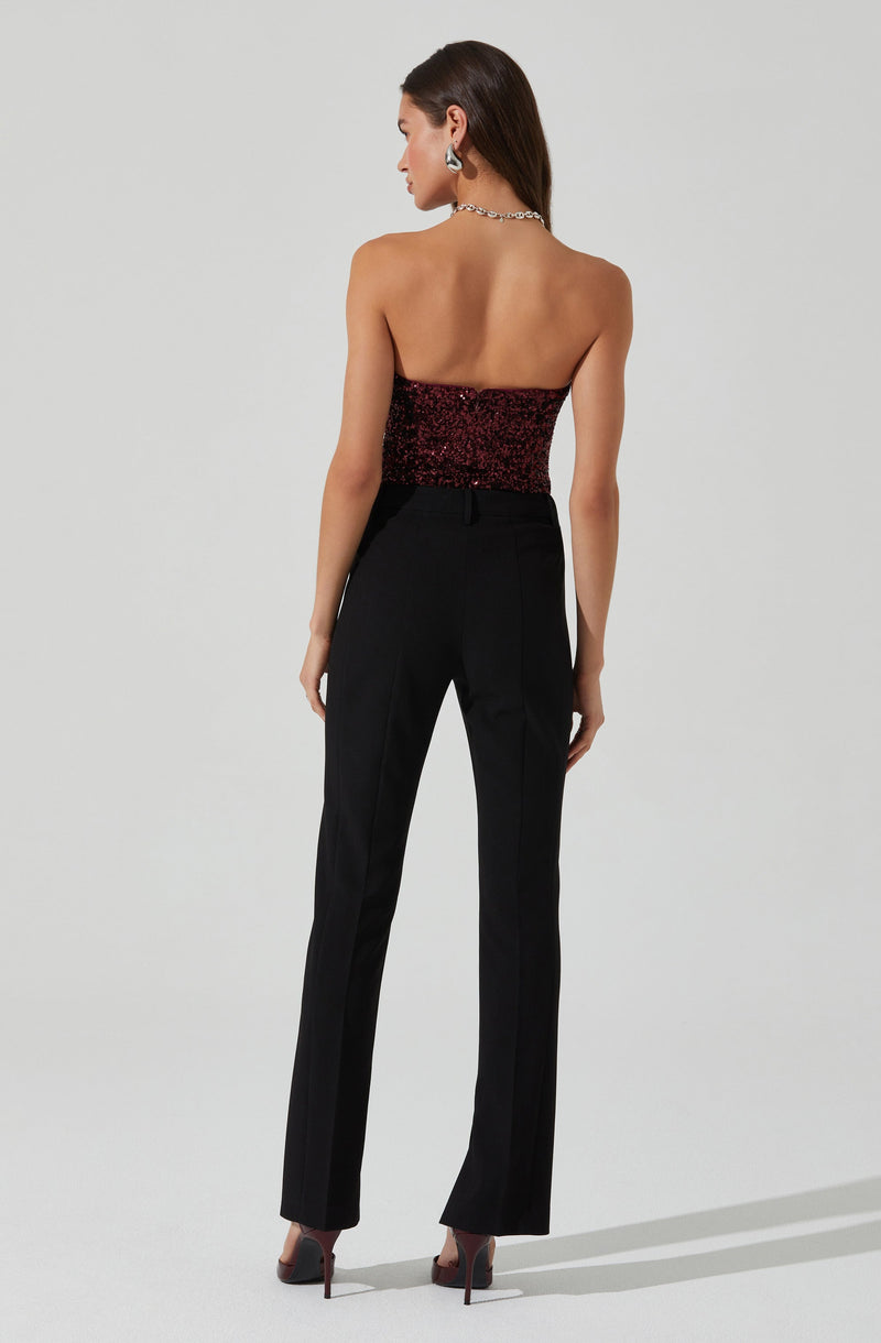 A woman is wearing a strapless, form-fitting top with a red and black sequined pattern, paired with high-waisted black trousers that have a tailored fit and slight flare at the bottom. She is also wearing high-heeled shoes, with her hair styled straight and accessorized with statement earrings and a necklace.