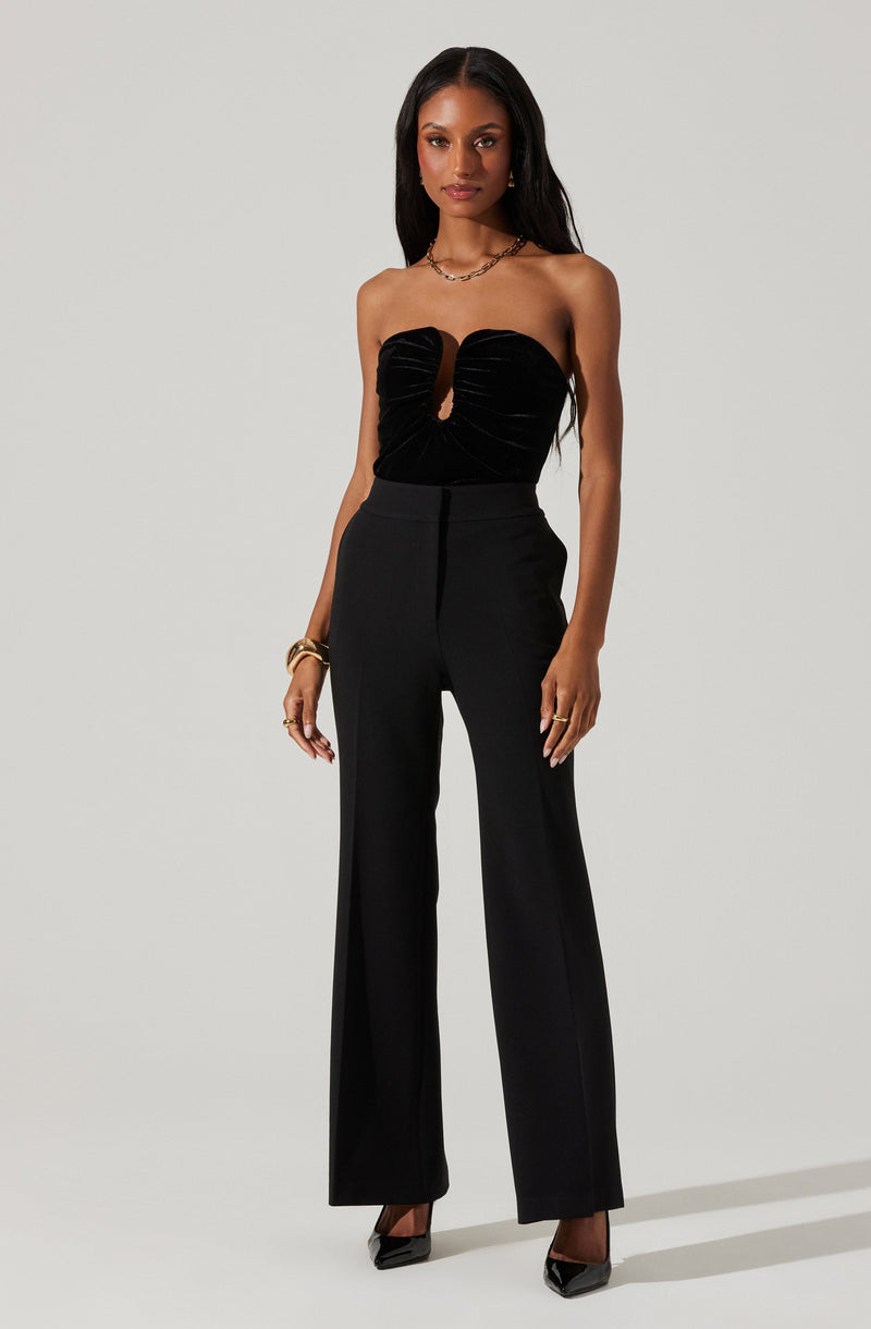 A woman is wearing a strapless black top with a keyhole cutout in the center, featuring gathered fabric that creates a ruched effect. She pairs it with high-waisted, wide-leg black trousers. Her accessories include a gold bracelet, multiple rings, and a necklace. She completes the look with black pointed-toe heels.