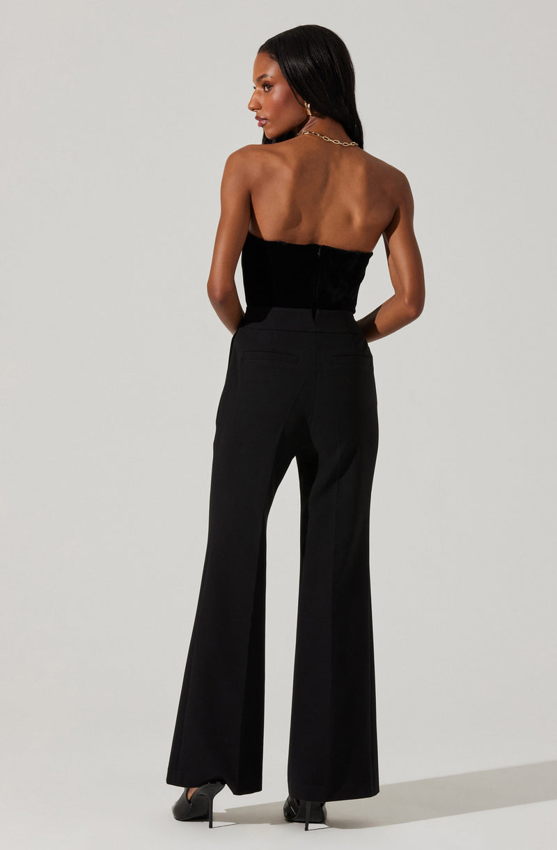 A woman is wearing a strapless black top made of a textured fabric, paired with high-waisted black wide-leg pants. She is also wearing black high heels, a gold chain necklace, and hoop earrings. Her hair is long and straight.