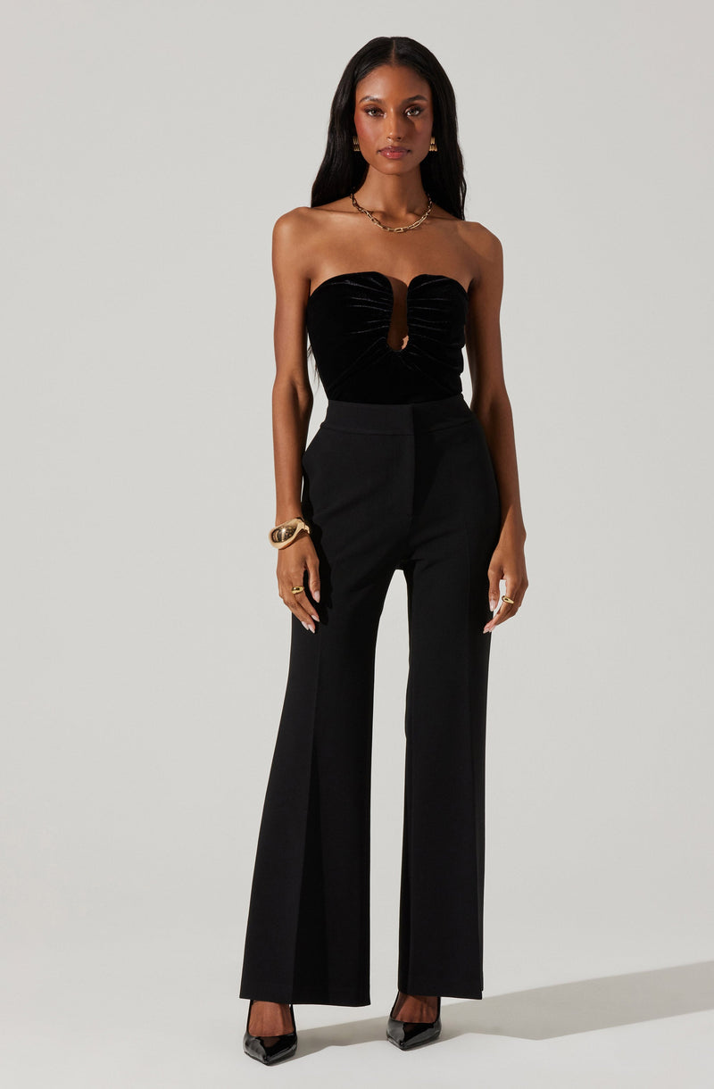 A woman is wearing a strapless black velvet top with a front keyhole detail, paired with high-waisted black wide-leg trousers. She accessorizes with gold jewelry, including a chunky necklace, bracelets, and rings, and is wearing black pointed-toe heels.