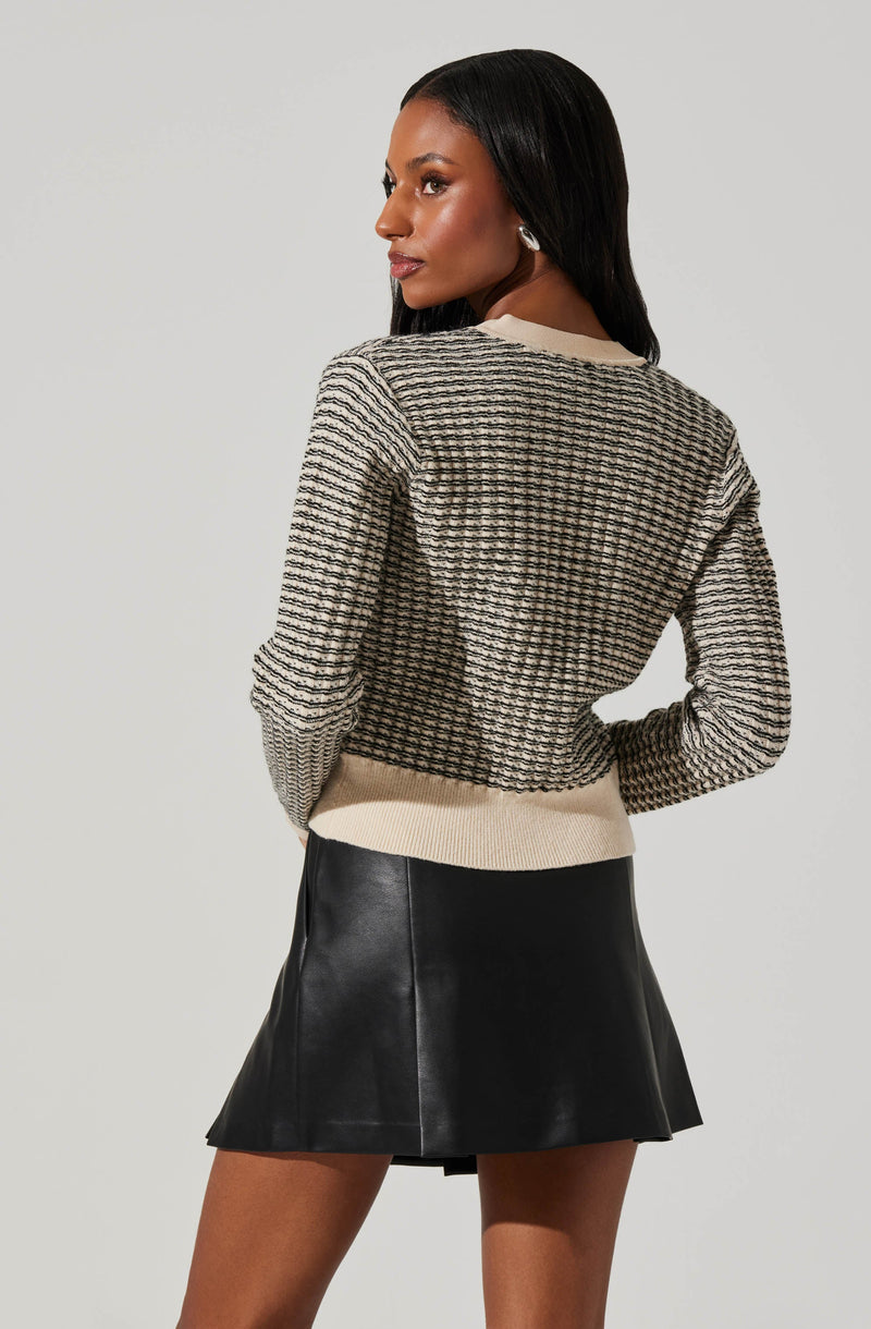 A woman is wearing a long-sleeved, knit sweater with a textured, checkered pattern in neutral tones. The sweater has a beige ribbed hem and collar. She is also wearing a black, leather-like A-line mini skirt.
