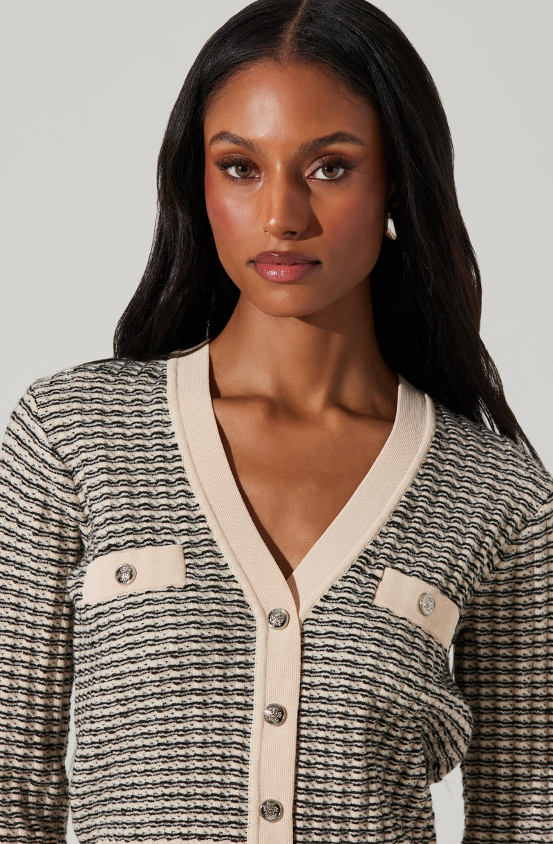 A woman is wearing a cream-colored cardigan with horizontal black and gray stripes. The cardigan features a V-neckline, cream-colored trim, and decorative buttons with diamond-like embellishments on the front pockets and center.