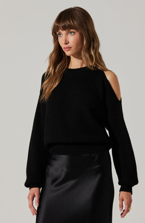 A woman is wearing a black, knitted sweater with shoulder cut-outs that reveal her skin. The sweater has a textured pattern and long sleeves with ribbed cuffs. She is also dressed in a black, shiny, satin or leather-like skirt that reaches to her knees.