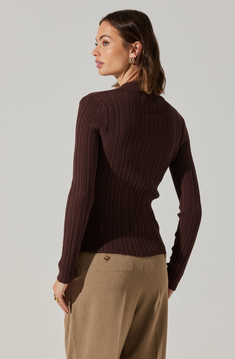 A woman is wearing a fitted, long-sleeved ribbed sweater in a dark brown color. The sweater has a crew neckline and vertical ribbing throughout. She is also dressed in beige trousers with a relaxed fit.