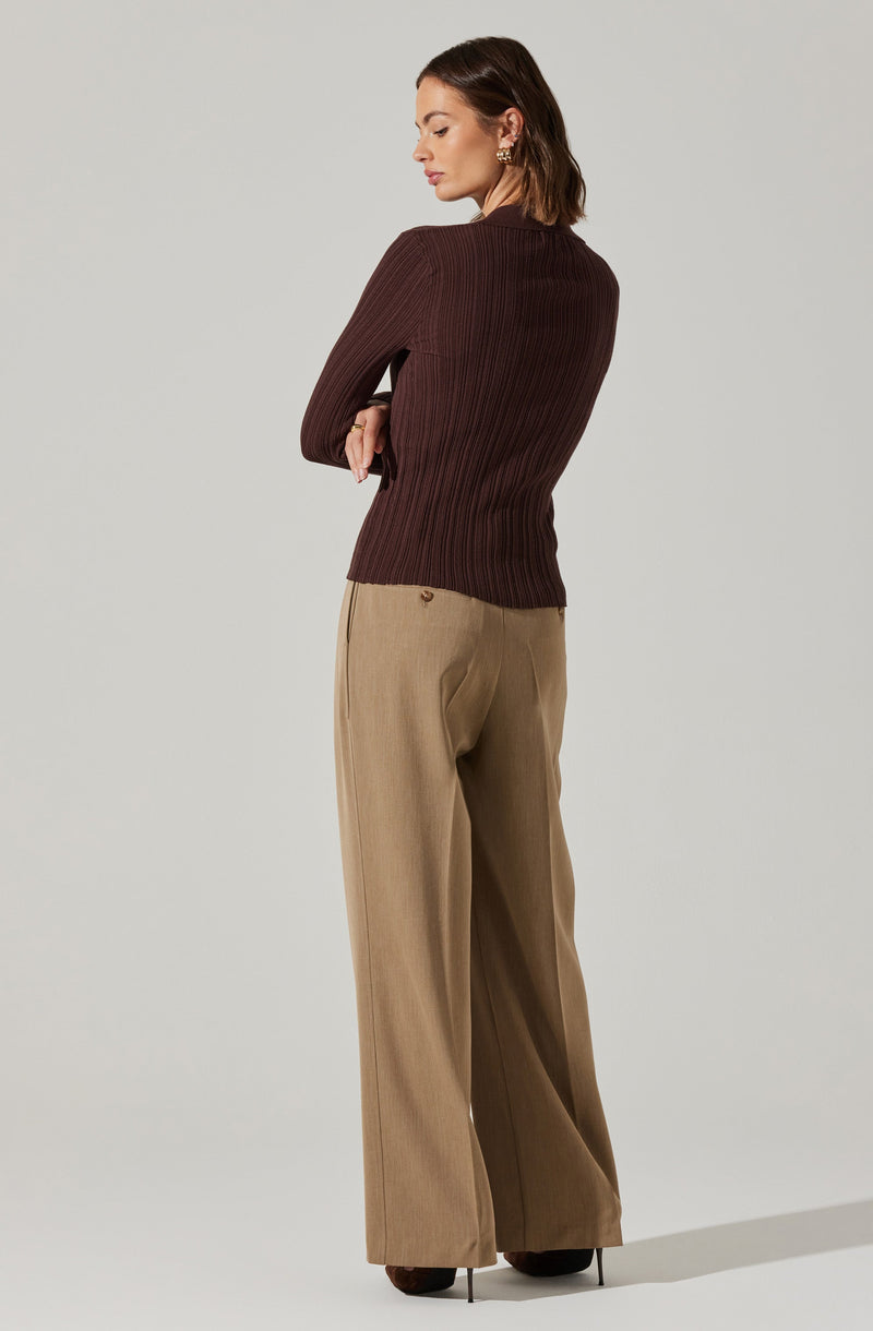 A woman is wearing a fitted, long-sleeved, ribbed sweater in a dark brown color and high-waisted, wide-leg beige trousers with a subtle pinstripe pattern. She has earrings and is standing in a neutral pose against a plain light background.