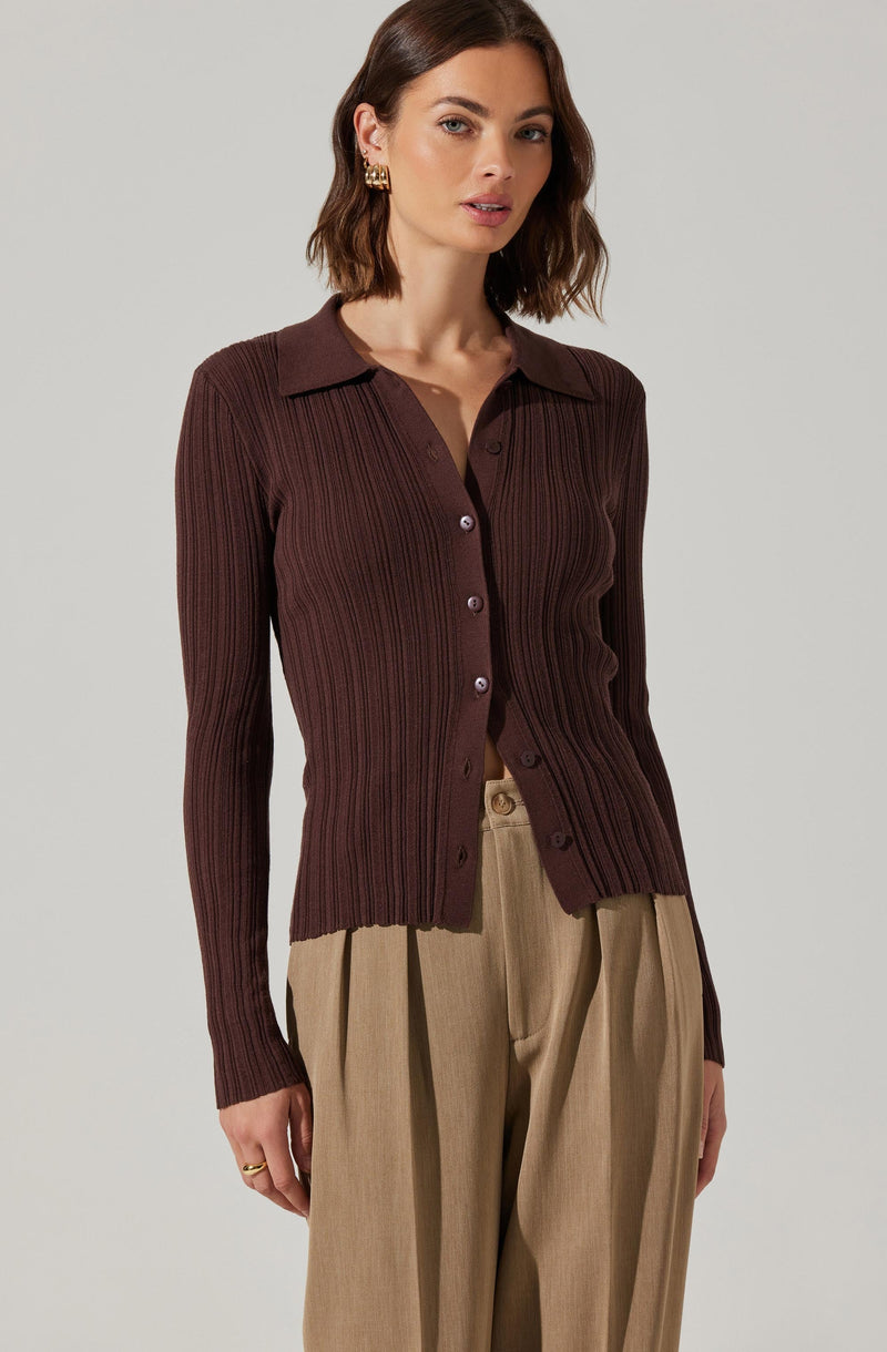 A woman is wearing a fitted, long-sleeved, dark brown ribbed knit cardigan with a collar and buttons down the front. The cardigan has vertical ribbing throughout and is styled with beige pleated trousers.