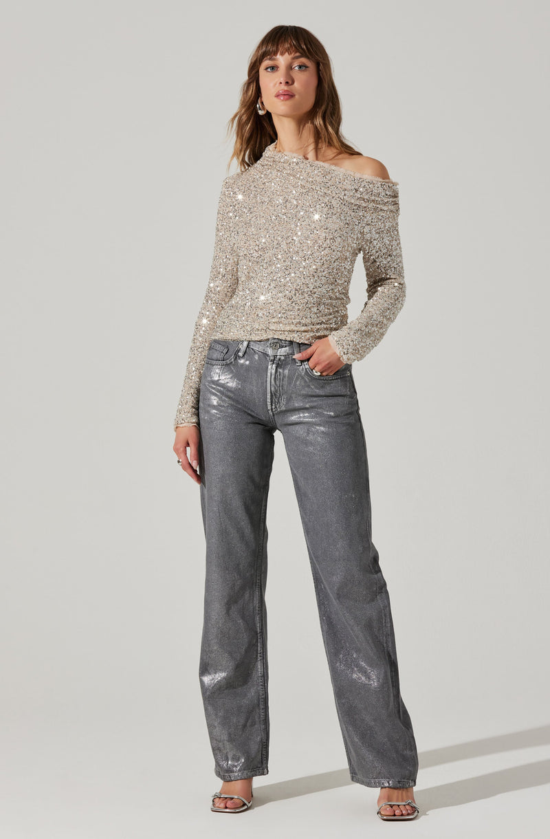 A woman is wearing an off-the-shoulder, long-sleeve, sparkly silver top with sequins, paired with high-waisted, straight-leg, metallic silver-grey jeans. She is also wearing open-toe heels.