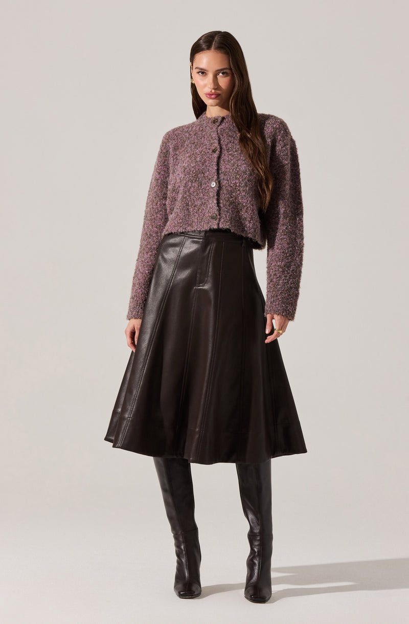 A woman is wearing a textured, long-sleeve, button-up cardigan in a mottled purple and gray color. She pairs it with a high-waisted, knee-length black leather flared skirt and black leather knee-high boots.