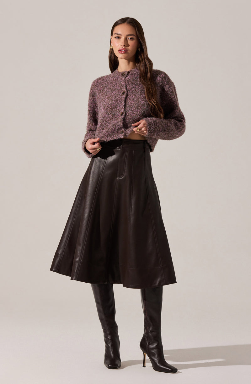 A woman is wearing a cropped, fuzzy, long-sleeved cardigan in shades of purple and gray with a button-up front. She pairs it with a high-waisted, dark brown leather A-line midi skirt and matching tall leather boots with high heels.